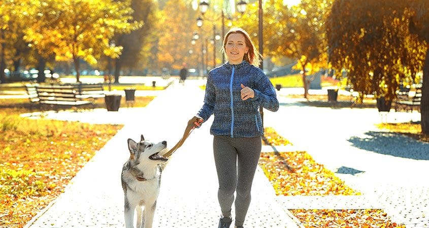 A woman runs with her dog on a fall day.